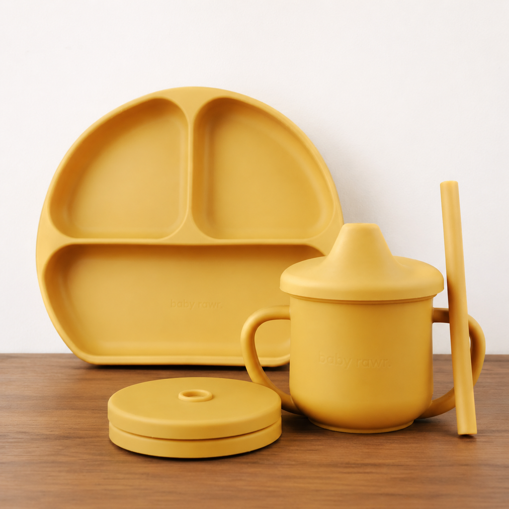 A yellow silicone baby feeding set on a wooden table against a white wall, including a three-compartment plate, a sippy cup with lid, a separate lid, and a chopstick.
